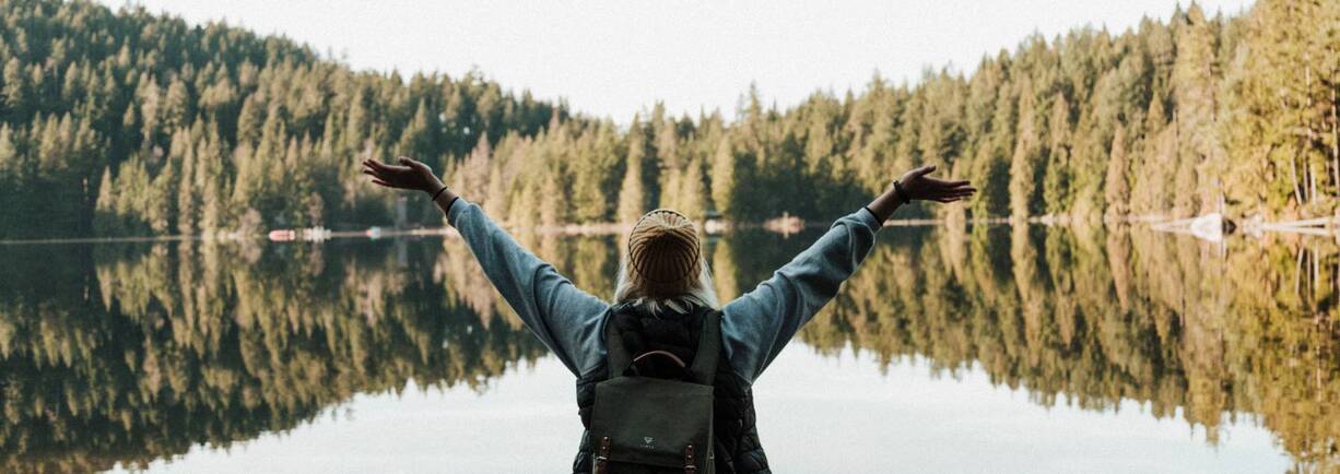 Eine junge Digitalnomadin steht vor einem See und freut sich.