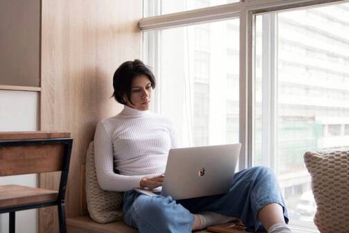 Eine junge Frau sitzt am Fenster mit ihrem MacBook und arbeitet. Eine digitale Nomadin, die ortsunabhängig arbeitet.