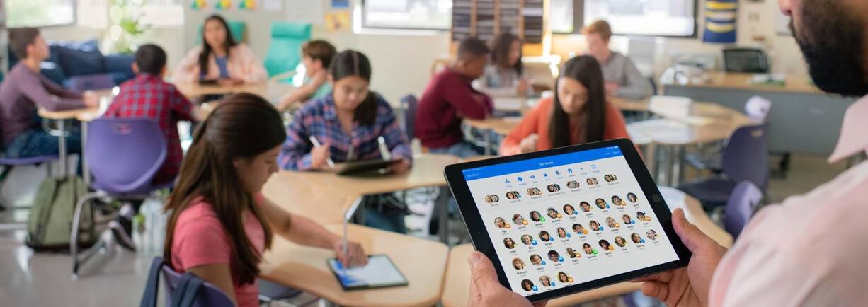 Lehrperson steht vor der Klasse mit iPad, das alle Lernenden zeigt