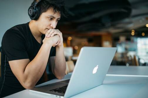 MacBook aufgeklappt und Mann davor Mann sitzt mit Kopfhörer vor seinem MacBook