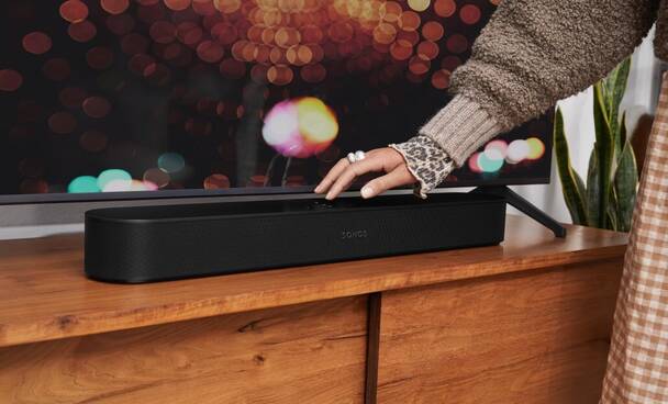 Schwarze Sonos Beam Soundbar unter TV-Gerät (Foto: Sonos) Frau bedient Touch-Feld der Sonos Beam (2. Gen) Soundbar