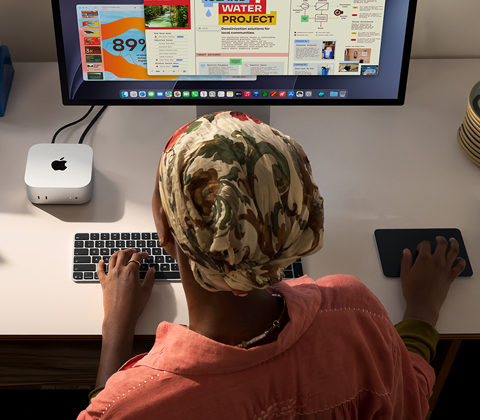 Eine Person tippt an einem Schreibtisch mit einem Mac mini, Studio Display angeschlossen, Magic Keyboard, Magic Trackpad, verschiedene Objekte