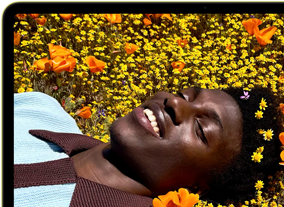 MacBook Neo, Farbe Zitrus, das Display zeigt eine Person, die in einer Blumenwiese in der Sonne liegt und lächelt