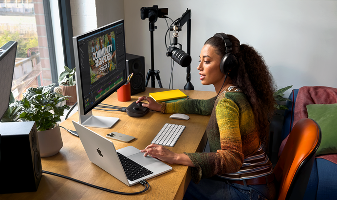 A woman in her home working at her desk with MacBook Pro connected to two external displays, wearing headphones and speaking into a microphone
