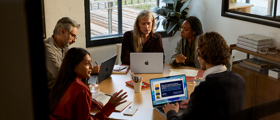 Eine Gruppe von fünf Mitarbeitenden sitzt an einem Tisch in einem Konferenzsaal, drei MacBook Pro Geräte werden bei dem Meeting verwendet.
