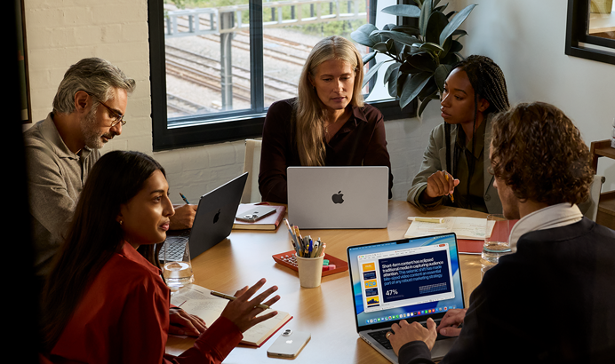 Eine Gruppe von fünf Mitarbeitenden sitzt an einem Tisch in einem Konferenzsaal, drei MacBook Pro Geräte werden bei dem Meeting verwendet.