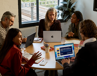 Eine Gruppe von fünf Mitarbeitenden sitzt an einem Tisch in einem Konferenzsaal, drei MacBook Pro Geräte werden bei dem Meeting verwendet.